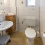 Badezimmer mit Toilette, Waschbecken und Spiegel. Holzschrank und Toilettenbürste sichtbar.