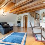Wohnzimmer mit Holzbalken, Sofa, Stühlen und Teppich. Treppe führt zur oberen Etage.