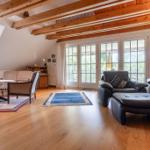 Wohnzimmer mit Holzbalken, großen Fenstern und Ledersofas auf Holzboden.
