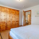 Schlafzimmer mit Holzschrank, Bett und Sessel. Tür führt in angrenzendes Zimmer.