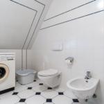 Badezimmer mit Waschmaschine, Toilette und Bidet. Weiße Fliesen mit dunklen Akzenten.