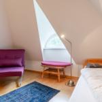 Schlafzimmer mit Bett, violettem Sessel und Fenster unter Dachschräge.