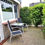 Terrasse mit Tisch und zwei Stühlen, umgeben von grünen Sträuchern und einem Haus.