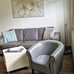 Ein Wohnzimmer mit grauem Sofa, Stuhl und Couchtisch. An der Wand hängt ein Bild mit Blumen.
