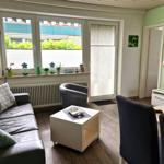 Wohnzimmer mit Sofa, Sessel, Tisch und TV. Fenster mit Pflanzen und Blick nach draußen.