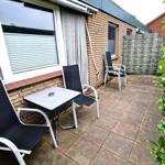 Terrasse mit Tisch und drei Stühlen, Wand mit Jalousie und Steinmauer im Hintergrund.