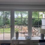 Durch große Fenster blickt man auf einen gepflegten Garten mit Holzstühlen und Steinmauer.