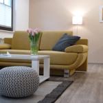 Gelber Sofa mit Kissen, weißer Couchtisch und grauer Hocker. Wandbild und Stehlampe.