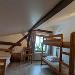 Zimmer mit zwei Etagenbetten und einem Einzelbett, Holzbalken an der Decke, Fenster mit Vorhängen.
