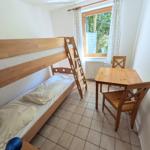 Zimmer mit Holzbett, Tisch und Stühlen. Fenster mit Vorhängen. Fliesenboden.
