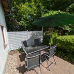 Terrasse mit Tisch, vier Stühlen und grünem Sonnenschirm. Umgeben von Bäumen und Hecke.