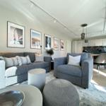 Wohnbereich mit grauem Sofa, Sessel und Wandbildern. Küche im Hintergrund.