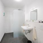 Badezimmer mit Dusche, Waschbecken und Spiegel. Weiße Fliesen und graue Bodenfliesen.