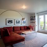 Ein Wohnzimmer mit rotem Sofa, Glas-Tisch und Bücherregal. Durchgang zur Terrasse mit Blick auf Grünflächen.