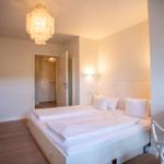 Schlafzimmer mit weißem Bett, Holzboden und Deckenleuchte. Tür führt in einen Flur.