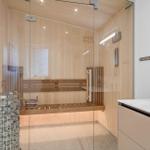 Sauna mit Holzwand, Sitzbank und Steinbehälter. Glaswand trennt Raum von Bad.