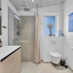 Badezimmer mit Dusche, Waschbecken und Toilette. Fenster und Wandbild mit Landschaft.