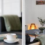 Tisch mit Tasse, Buch und Pflanze. Hintergrund: Sofa mit Decke und Wandbild.