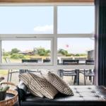 Ein schwarzes Sofa mit Kissen vor großen Fenstern mit Blick auf eine Terrasse und Landschaft.