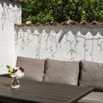 Außenbereich mit Sitzbank, Tisch und Blumenarrangement vor weißer Wand mit Pflanzen.