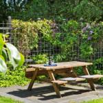 Holzpicknicktisch mit Flasche und Gläsern auf Steinplatten. Hintergrund: Ranken mit lila Blüten an Zaun.