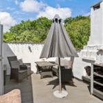Terrasse mit Sitzgruppe, Sonnenschirm und Grillbereich. Weiße Mauer und grüne Bäume im Hintergrund.
