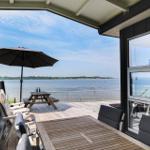 Terrasse mit Tisch, Stühlen und Sonnenschirm. Blick auf ruhiges Wasser und gegenüberliegende Ufer.