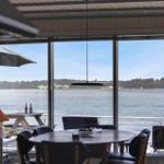 Glasfront mit Blick auf Wasser und Ufer, Tisch mit Stühlen und Terrasse mit Sonnenschirm.