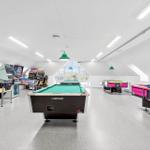Raum mit Billardtischen und Spielautomaten. Grüne Deckenleuchten und große Fenster.