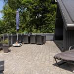 Terrasse mit Liegestuhl, Tisch, Stühlen und Sonnenschirm. Hintergrund: grüne Bäume und Holzzaun.