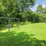 Großer Garten mit Schaukelanlage und grünem Rasenbereich. Umgeben von Bäumen und Sträuchern.