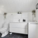 Badezimmer mit Toilette, Waschbecken und Waschmaschine. Weiße Wand- und Deckenverkleidung.