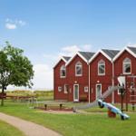 Rote Holzhäuser mit Spielplatz und Grünfläche. Strand im Hintergrund.