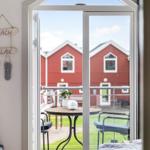 Durch die Glastür blickt man auf eine Terrasse mit Tisch und Stühlen. Im Hintergrund sind rote Häuser zu sehen.