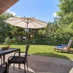 Terrasse mit Tisch, Stühlen und Sonnenschirm. Hintergrund: grüner Garten mit Liegestühlen.