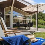 Terrasse mit Liegestühlen, Sonnenschirm und Tisch. Hintergrund: Haus mit Glasfront und Grün.