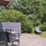 Terrasse mit Tisch, Stühlen und Sonnenschutz. Hinterhof mit Liegestuhl und Holzstapel.