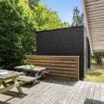 Holzterrasse mit Picknicktisch, Sitzbank und Blick auf einen schwarzen Holzschuppen.