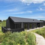Moderne schwarze Hütte mit Holzterrasse und Weg durch Grasland unter blauem Himmel.