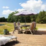 Holzterrasse mit Tisch, Stühlen und Sonnenschirm. Roter Holzschuppen und Grünfläche im Hintergrund.