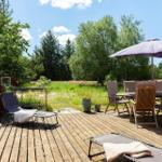 Holzterrasse mit Liegestühlen, Tisch und Sonnenschirm. Grünflächen und Bäume im Hintergrund.