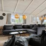 Wohnzimmer mit grauem Sofa, Tisch und Sitzgelegenheiten. Holzwände und Decke. Fenster mit Blick nach draußen.