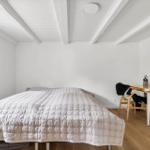 Schlafzimmer mit Bett, Schreibtisch und Fenster. Holzfußboden und weiße Wände.