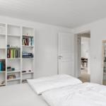 Schlafzimmer mit Bett, Bücherregal und Spiegel. Tür führt in einen weiteren Raum.