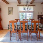 Esszimmer mit Holztisch, Stühlen, Tischdecke und Kronleuchter. An der Wand hängen Bilder und ein großer Wecker.