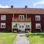 Rotes Holzhaus mit Balkon, weißen Fenstern und Eingang, umgeben von Gras und Bäumen.