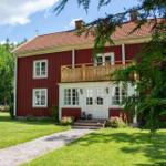 Rotes Haus mit Balkon, weißen Fenstern und Eingangstür. Grünflächen und Bäume umgeben das Gebäude.