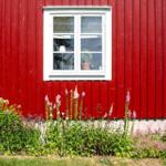 Rotes Holzhaus mit weißem Fenster und Blumenbeet im Vorgarten.