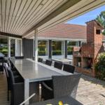 Terrasse mit Tisch, Stühlen, Grill und Backofen. Hintergrund: Haus und Bäume.