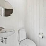 Badezimmer mit Waschbecken, Toilette und runden Spiegel an weißen Holzwänden.